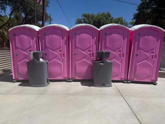 Five pink porta potties lined up with two gray handwashing stations on concrete