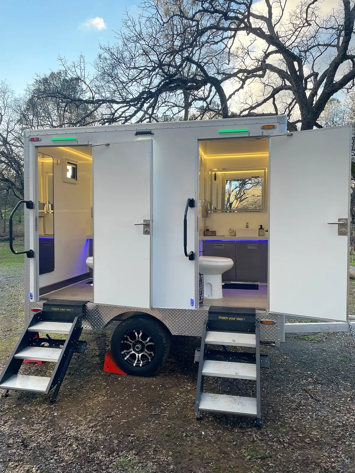 Restroom trailer with both doors open and interior lights glowing in the evening