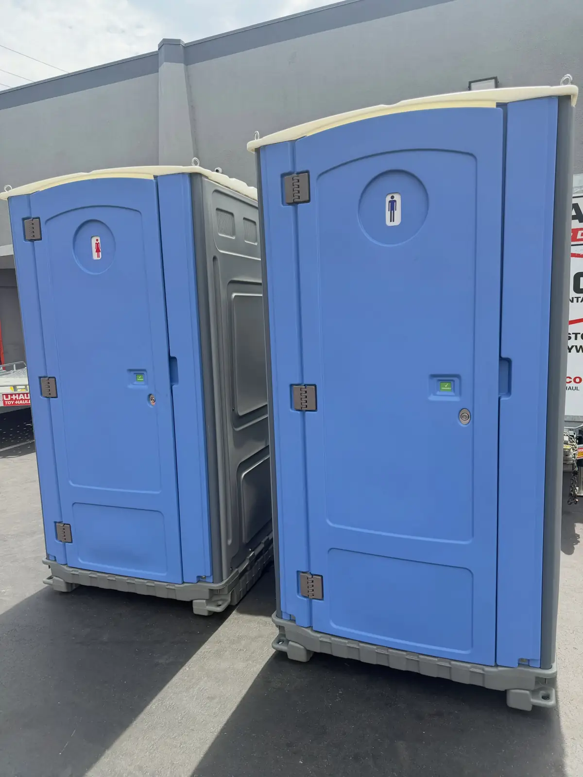 Two porta potties side by side on a paved lot next to a building