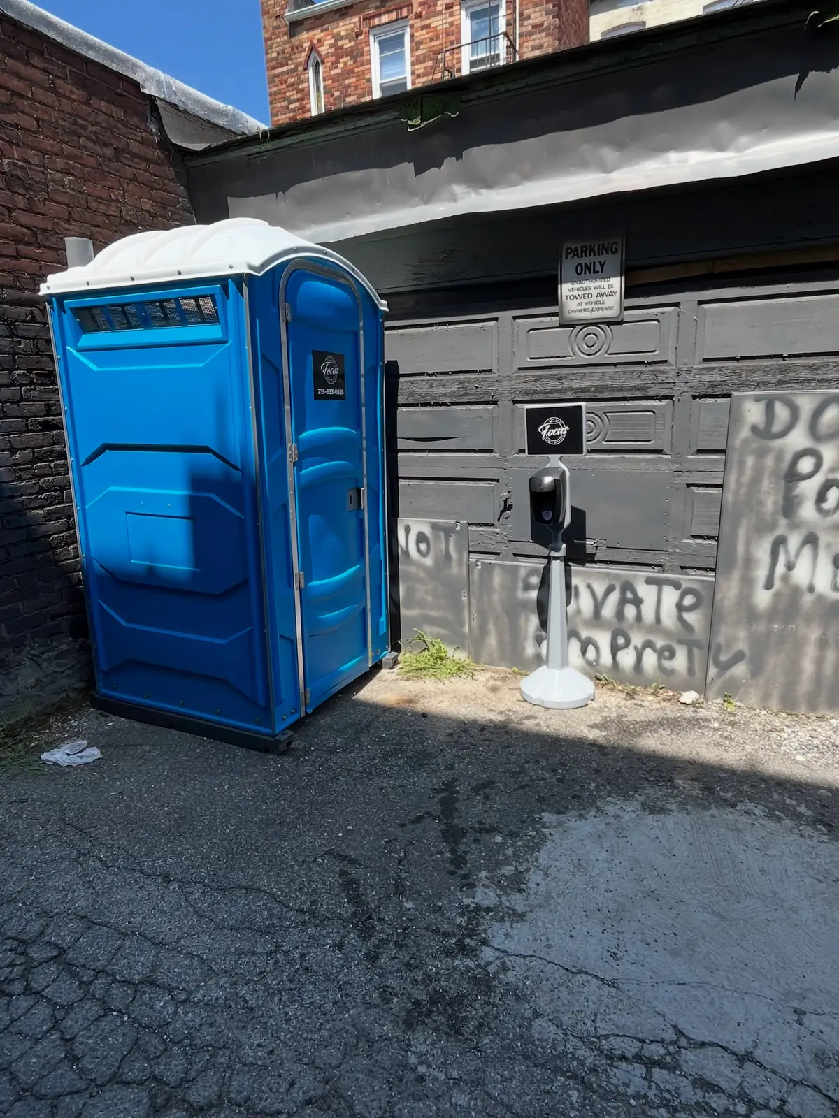 Blue standard porta potty placed in a paved alley behind a brick building