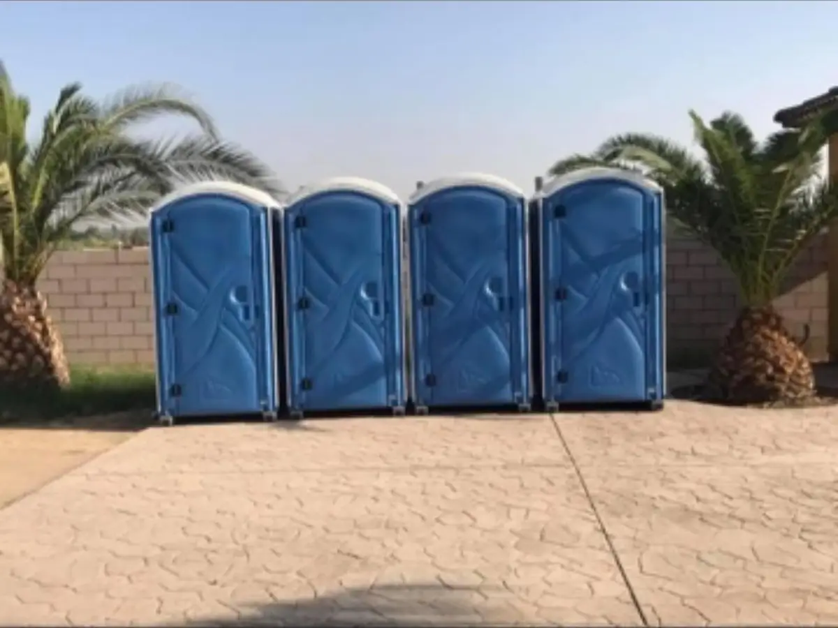 Four blue standard porta potties standing in a row along a paved driveway