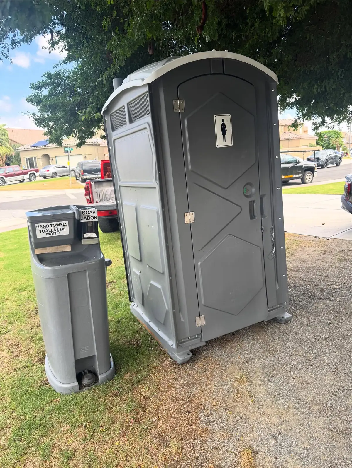 Gray standard porta potty beside a gray foot-pump handwashing station on grass